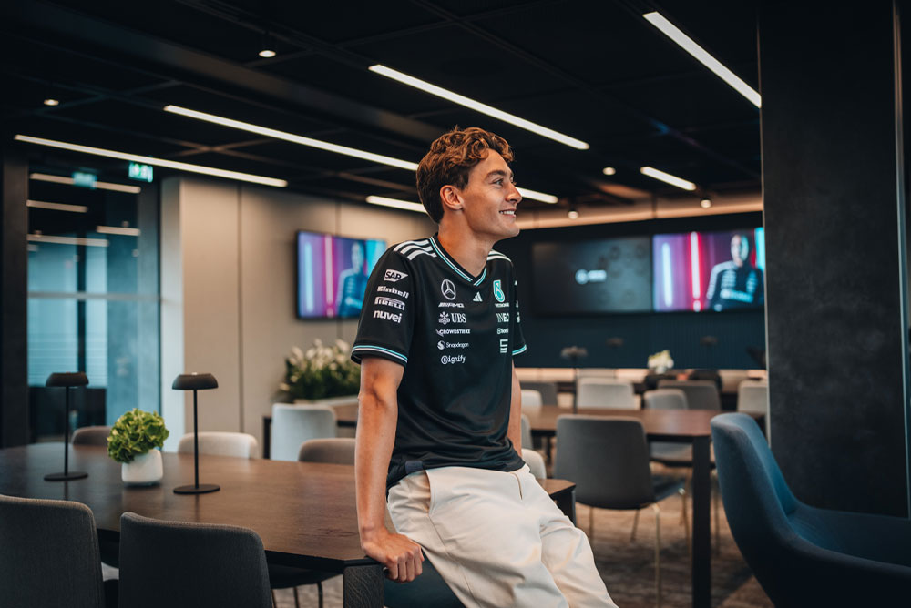 George Russell, pilota del team Mercedes-AMG PETRONAS Formula One Team, è seduto in modo rilassato su un tavolo in un’area hospitality o ufficio del team, moderna ed elegante. Sorride e indossa la maglietta ufficiale del team con i loghi degli sponsor visibili, come Einhell, IWC e CrowdStrike. Sugli schermi sullo sfondo appare la sua immagine.
