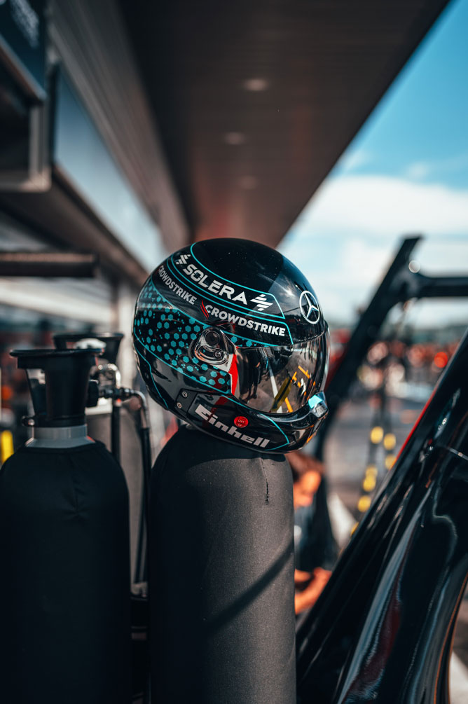 Un casco del Mercedes-AMG PETRONAS Formula One Team con i loghi ben visibili degli sponsor, tra cui Einhell, CrowdStrike e Solera, è posato su una bombola del gas nella corsia dei box. Il riflesso sulla visiera mostra i dintorni del garage.