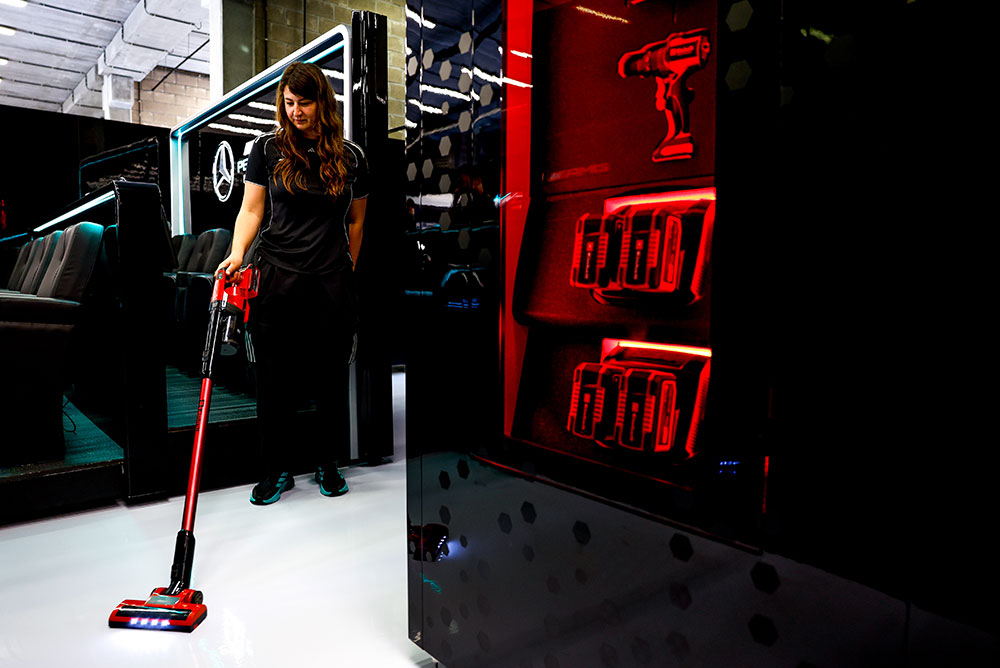 Una donna del Mercedes-AMG PETRONAS Formula One Team sta pulendo un pavimento bianco lucido con un aspirapolvere senza fili Einhell. A destra, scaffali illuminati di rosso espongono diverse batterie Einhell Power X-Change e un trapano a batteria.