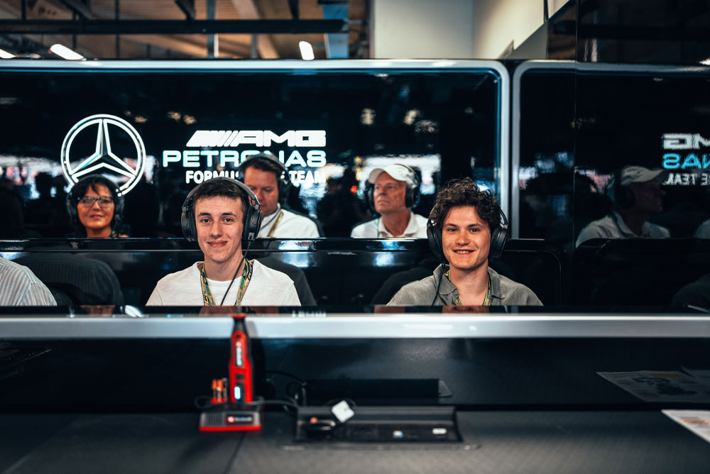 Due uomini sono seduti nel garage del Mercedes-AMG PETRONAS Formula One Team, indossano cuffie e si godono l’esperienza dietro le quinte. In primo piano è visibile un dispositivo Einhell rosso.