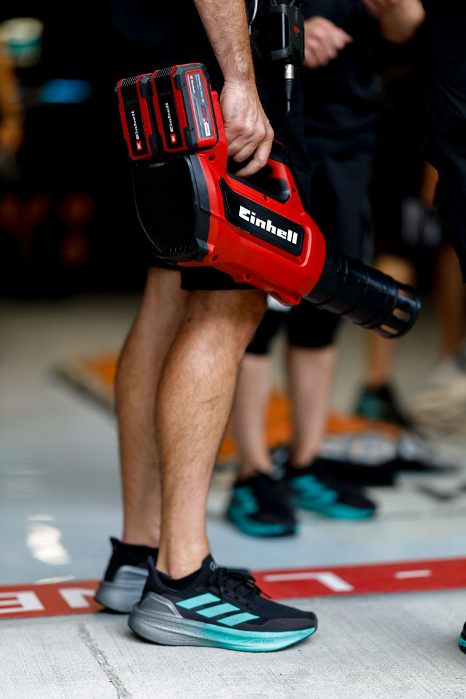 Un membro del team tiene in mano un soffiatore senza fili Einhell rosso con due batterie Power X-Change. La scena si svolge nella zona dei box durante un evento di Formula 1.