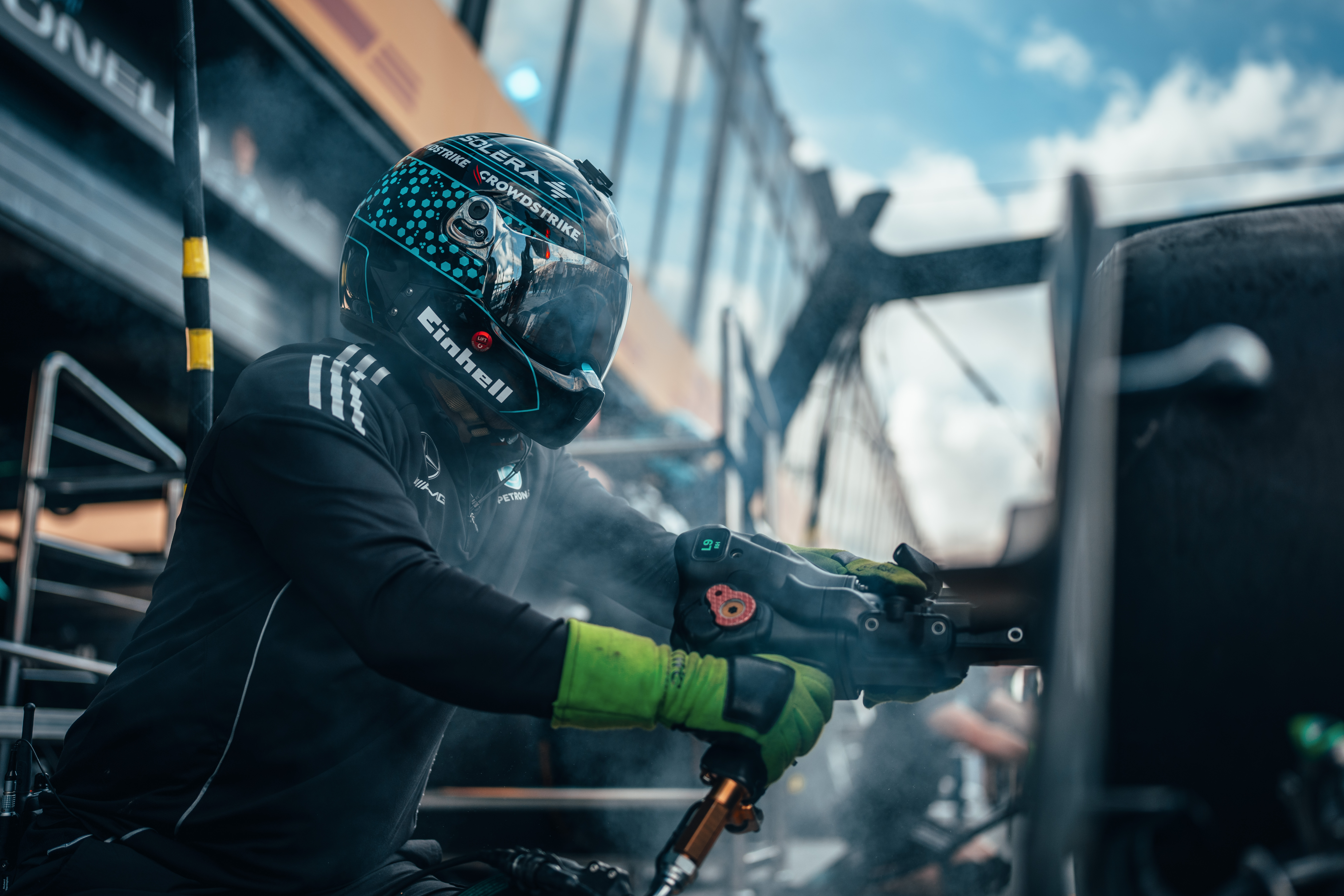 Un membro della squadra dei box di Formula 1, con casco e guanti, utilizza una pistola pneumatica per le ruote durante un pit stop, circondato da fumo e attrezzature.