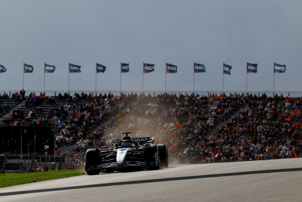 Un’auto del Mercedes-AMG PETRONAS Formula One Team passa davanti a tribune gremite durante una gara. I tifosi sventolano bandiere sullo sfondo, creando un’atmosfera vibrante.