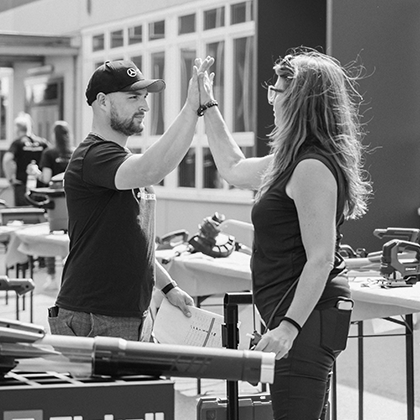 Un uomo e una donna si danno il cinque durante un evento Einhell all'aperto con esposizione di utensili, in bianco e nero.