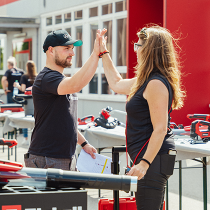 Un uomo e una donna si danno il cinque durante un evento Einhell all'aperto con esposizione di utensili, mostrato a colori.