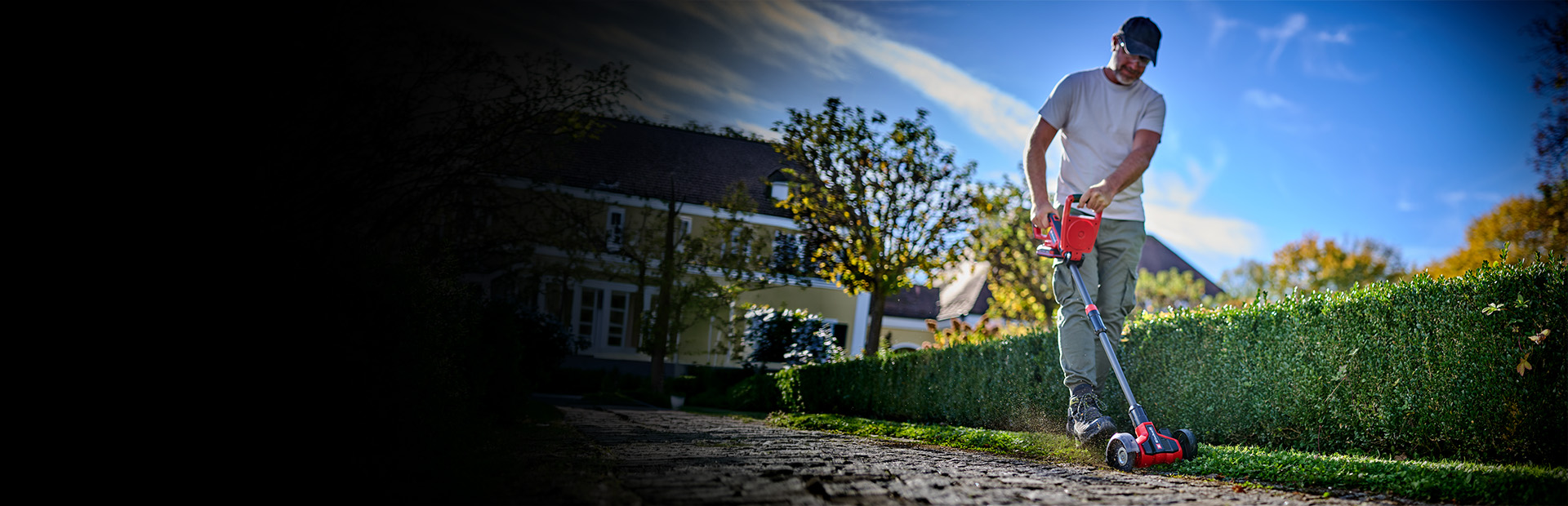 A man cleans paving joints along a hedge in the garden using the Einhell cordless patio cleaner.